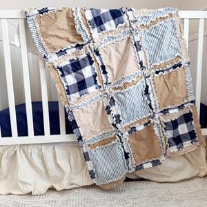 May include: A handmade patchwork quilt with a mix of fabrics in tan, navy blue gingham, and blue and white stripes. The quilt is draped over a white crib, showcasing its textured design and cozy aesthetic. The quilt's dimensions are not visible.