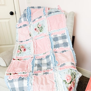 May include: A handmade patchwork quilt in shades of pink, blue, and gray, draped over a chair. The quilt features a mix of floral, gingham, and solid fabric squares, with ruffled edges. The overall design is a cozy, cottagecore aesthetic.