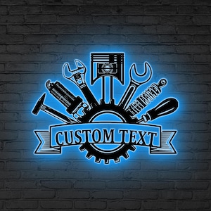 Peut inclure: Enseigne au néon noire et bleue représentant des outils de mécanicien, dont des clés et un piston, disposés autour d'un engrenage. Le texte "CUSTOM TEXT" est inscrit sur une bannière.