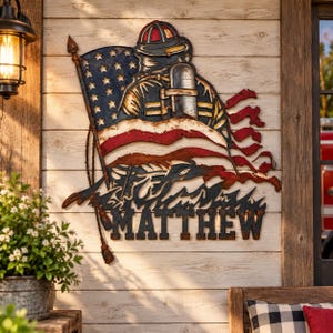 May include: A metal wall art piece featuring a firefighter in a helmet and gear standing before a waving American flag. The design includes a rope detail on the left side and the name MATTHEW in bold, dark, distressed-finish capital letters.