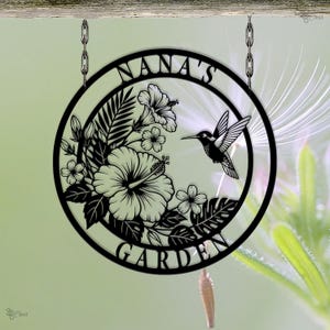 Może przedstawiać: Czarny metalowy znak ogrodowy z napisem "NANA'S GARDEN" w okrągłym kształcie. Znak przedstawia kolibra, kwiaty hibiskusa i tropikalne liście. Znak wisi na drewnianej belce z metalowymi łańcuchami.