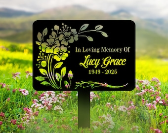 May include: A black memorial garden stake with floral accents and the text "In Loving Memory Of Lucy Grace 1949-2025." The flowers are yellow and silver, with green leaves. The stake is in a garden setting.