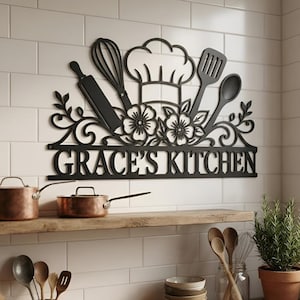 以下が含まれることがあります： GRACE'S KITCHENという文字が刻まれた黒い金属製の壁掛けサイン。シェフハット、泡立て器、麺棒、ヘラ、スプーンのモチーフが配置され、花や蔓の装飾的な切り抜きが施されています。
