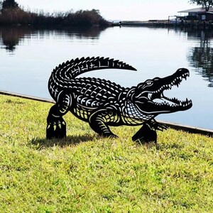 May include: A black metal alligator sculpture with open jaws, positioned on a grassy bank near water. The sculpture features detailed cut-out patterns, depicting the alligator's scales and form. The background shows a lake and trees.