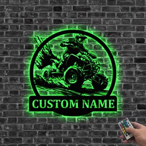 Könnte beinhalten: Schwarzes Metallschild mit ATV-Motiv und neongrüner Beleuchtung, das einen Fahrer auf einem Geländefahrzeug zeigt. Das Schild ist an einer Backsteinmauer angebracht und enthält den Text "CUSTOM NAME". Eine Fernbedienung ist unten rechts sichtbar.