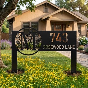 Puede incluir: Un letrero de dirección de casa de metal negro con un recorte circular que presenta una letra M decorativa rodeada de motivos de rosas. El letrero muestra el número 743 y el texto ROSEWOOD LANE en una fuente moderna, sobre dos soportes.
