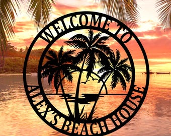 Könnte beinhalten: Schwarzes Metallschild mit einer Strandszene. Das Schild zeigt Palmen, das Meer und den Text "WELCOME TO ALEX'S BEACH HOUSE" in einem kreisförmigen Design. Der Hintergrund zeigt einen Sonnenuntergang über dem Wasser.