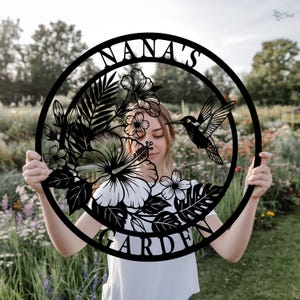 Może przedstawiać: Czarny metalowy znak ogrodowy z napisem "NANA'S GARDEN" w okrągłym kształcie. Znak przedstawia kolibra, kwiaty i liście. Znak jest trzymany przed ogrodem.