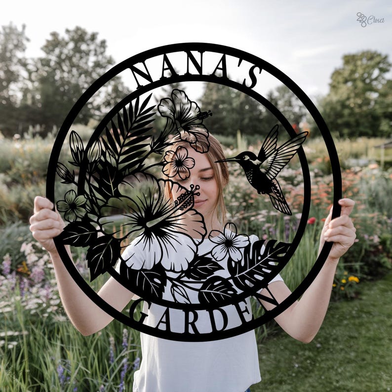 Może przedstawiać: Czarny metalowy znak ogrodowy z napisem "NANA'S GARDEN" w okrągłym kształcie. Znak przedstawia kolibra, kwiaty i liście. Znak jest trzymany przed ogrodem.