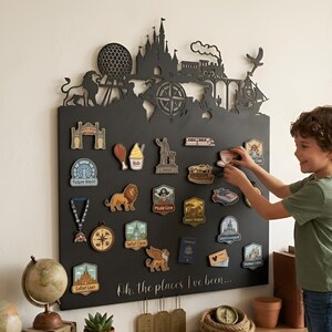 Puede incluir: Un tablero de pared rectangular negro con una silueta cortada con láser de un castillo, un tren y una brújula en la parte superior. El tablero incluye el texto "Oh, the places I've been..." y está decorado con varios imanes coloridos.