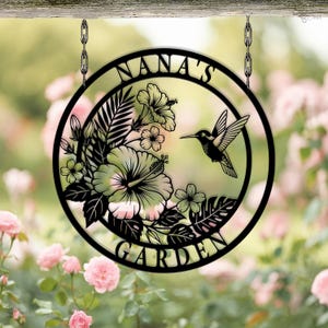 Może przedstawiać: Czarny metalowy znak ogrodowy z napisem "NANA'S GARDEN" w okrągłym kształcie. Znak przedstawia kolibra, kwiaty hibiskusa i tropikalne liście. Znak wisi na dw&oacute;ch łańcuchach.