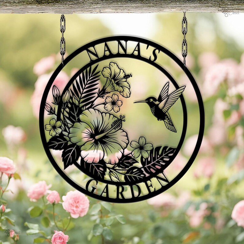 Może przedstawiać: Czarny metalowy znak ogrodowy z napisem "NANA'S GARDEN" w okrągłym kształcie. Znak przedstawia kolibra, kwiaty hibiskusa i tropikalne liście. Znak wisi na dw&oacute;ch łańcuchach.