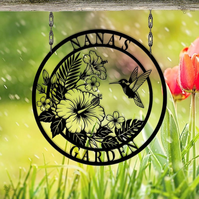 Może przedstawiać: Czarny metalowy znak ogrodowy z napisem "NANA'S GARDEN" w okrągłym kształcie. Znak przedstawia kolibra, kwiaty hibiskusa i tropikalne liście, wiszące na łańcuchach.