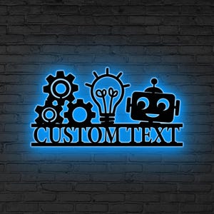 Könnte beinhalten: Schwarze Metall-Wandkunst mit Zahnrädern, einer Glühbirne und einem Roboter-Design. Die Worte "CUSTOM TEXT" sind unter dem Design zu sehen. Die Kunst wird von einem blauen Neonlicht beleuchtet und an einer Backsteinmauer befestigt.