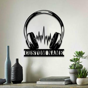 May include: Black metal wall art featuring a pair of headphones with a sound wave design and a customizable nameplate. The nameplate reads "CUSTOM NAME".