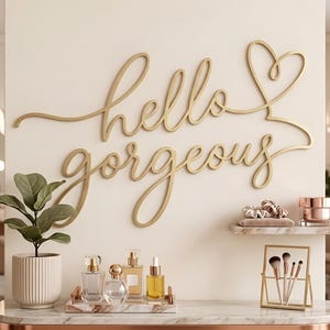May include: A gold-coloured wall sign featuring the cursive text hello gorgeous with a heart shape at the end. The sign is mounted on a light-coloured wall above a vanity area containing a potted plant, perfume bottles, and makeup brushes.
