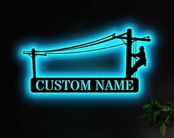 May include: Black metal wall art featuring a lineman climbing a utility pole, with a glowing blue outline. The text "CUSTOM NAME" is displayed on a rectangular sign below the pole. The artwork is mounted on a dark wall.