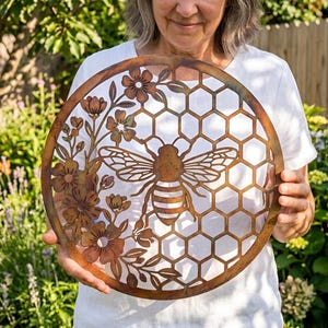 May include: A circular metal wall art piece featuring a rust-coloured finish. The design includes a central bee silhouette surrounded by a honeycomb pattern on the right side and a cluster of flowers and leaves on the left side of the frame.