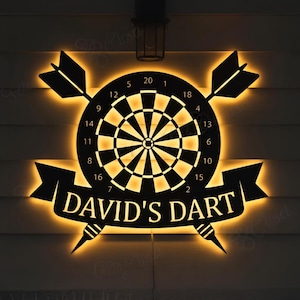 May include: A black and gold illuminated dartboard sign with the text "DAVID'S DART". The sign features a dartboard with two crossed darts and two additional darts. The sign is lit with a warm, inviting glow.