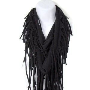 May include: A black fringe scarf made of soft fabric. The scarf is folded in a loop and has long fringe on all sides.