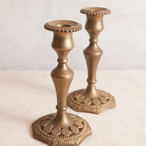 May include: Two antique brass candlesticks. The candlesticks have a detailed, ornate design with a round base and a fluted top. The brass has a warm, aged patina, adding to their vintage charm. The candlesticks are a decorative home decor item.
