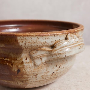 May include: A handmade ceramic bowl with a speckled, off-white exterior and a rich brown interior. The bowl features two decorative handles with a leaf-like design. The rustic pottery is ideal for serving soups, stews, or salads.