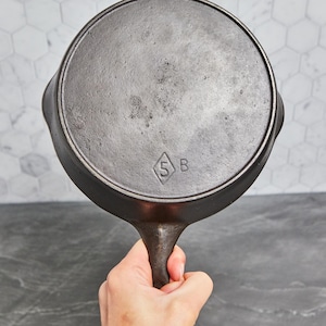 May include: A well-worn, cast iron skillet, held by a hand, with a long handle and two small side handles. The bottom of the pan is visible, showing a diamond-shaped marking with the numbers "5" and the letter "B".