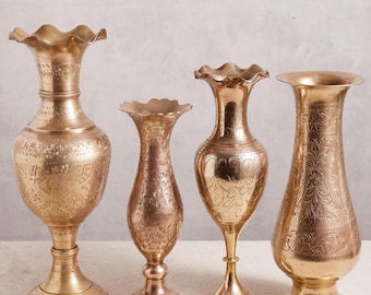 May include: Four antique brass vases with detailed floral designs and diverse shapes. The vases feature ruffled rims and pedestal bases. The brass has a warm, golden tone, and the vases are arranged on a light surface against a neutral backdrop.