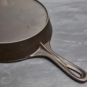 May include: A well-worn, dark brown cast iron skillet with a long handle and a circular cooking surface. The skillet has a number 7 marking on the bottom. The handle features a hole for hanging.