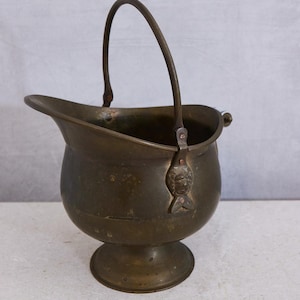 May include: A brass coal scuttle with a rounded body, a pedestal base, and a curved handle. The vessel features a wide, open spout and decorative metal attachments where the handle connects to the sides, showcasing a weathered, dark metallic finish.