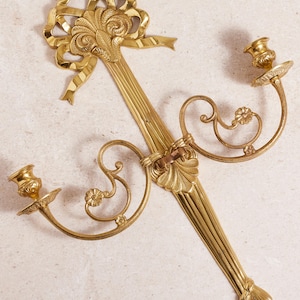 May include: A gold-colored, ornate wall sconce with two candle holders. The sconce features a decorative bow and floral accents. The design is symmetrical, with a central column and curved arms holding the candle cups. The overall style is vintage.
