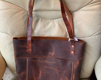 May include: A brown leather tote bag with a front pocket and two straps. The bag has a distressed look and is made of high-quality leather.
