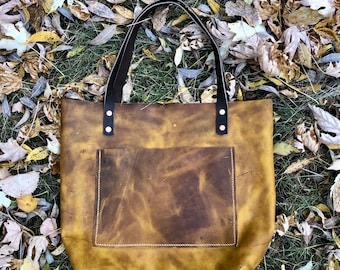 May include: A handcrafted, golden-brown leather tote bag with a front pocket and black handles. The bag is set against a backdrop of fallen leaves and grass, showcasing its rustic aesthetic. The handles are attached with rivets.