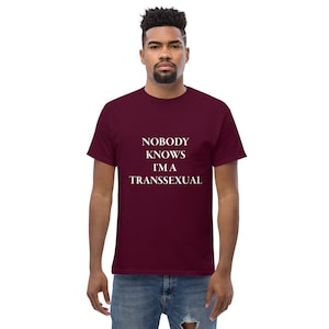 Puede incluir: Una camiseta color granate con el texto "NOBODY KNOWS I'M A TRANSEXUAL" impreso en blanco.