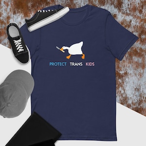 以下が含まれることがあります： 白いガチョウがくちばしでナイフを持っている、ネイビーブルーのTシャツ。ガチョウの下には「PROTECT TRANS KIDS」という文字が青、ピンク、白で印刷されています。