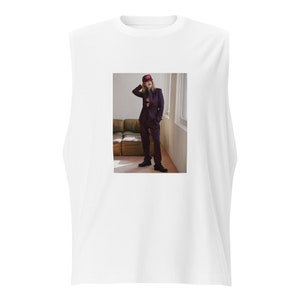 Op de afbeelding: Een wit, mouwloos T-shirt met een foto van een persoon die een paars pak en een roze baseballpet draagt. De persoon staat in een kamer met een bank op de achtergrond.