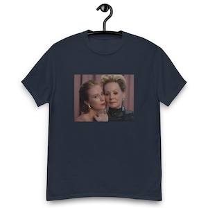 Op de afbeelding: Marineblauw T-shirt met een ingelijste afbeelding van twee vrouwen. De afbeelding toont twee vrouwen, een met blond haar en de andere met rood haar, die dicht bij elkaar poseren. Het t-shirt hangt aan een zwarte hanger.