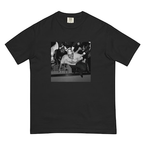 May include: Black t-shirt with a black and white photograph of a person jumping. The image shows a person in a light-coloured shirt and hat, surrounded by others. The t-shirt has a classic crew neck and short sleeves.