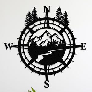 May include: Black metal wall art featuring a compass design with mountain and river silhouettes. The compass points North, South, East, and West. Two trees are at the top of the compass. The art is mounted on a white wall above a planter with green plants.