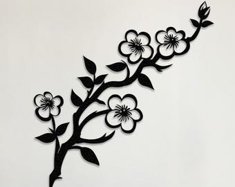 May include: Black metal wall art featuring a stylized branch with five white flowers. The branch is curved and has a delicate, flowing design.