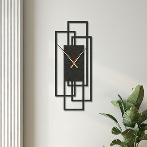 Puede incluir: Un reloj de pared de metal negro con manecillas doradas y fondo negro. El reloj está rodeado por un marco geométrico de cuadrados superpuestos. El reloj está colgado en una pared blanca con una planta en una maceta delante de él.