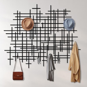 Könnte beinhalten: Ein moderner Wandorganizer mit einem schwarzen geometrischen Design. Er verfügt über Haken zum Aufhängen von Gegenständen wie Hüten, Mänteln, einer Handtasche und Schlüsseln. Zwei Hüte, ein beiger und ein blauer, werden zusammen mit einem beigen Mantel und einem grauen Schal gezeigt.