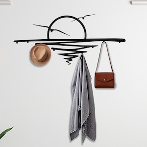 May include: Black metal wall-mounted coat rack with a stylised sunset design. The rack has three hooks and is decorated with a silhouette of a sun and birds. A brown hat, a grey scarf, and a brown purse are hanging on the hooks.