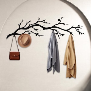 Op de afbeelding: Zwarte metalen wandbevestigde kapstok in de vorm van een tak met drie haken. Een hoed, een handtas en twee jassen hangen aan de haken.