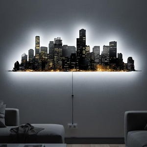May include: A black and gold cityscape wall art with a bright white LED backlight. The art piece depicts a silhouette of a city skyline with illuminated windows. The artwork is mounted on a white wall in a modern living room setting.