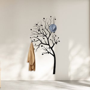 May include: A black silhouette of a tree with branches extending out, creating a wall-mounted coat rack. A brown jacket hangs from one of the branches, and a blue hat hangs from another branch.
