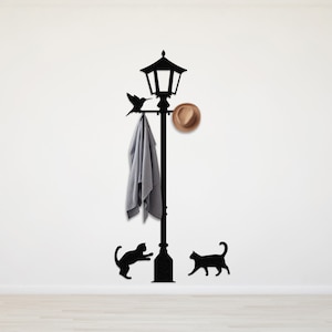 May include: Black silhouette of a street lamp with a bird perched on the arm, a hat hanging from the hook, and a jacket hanging from the hook. Two black cats are at the base of the lamp.