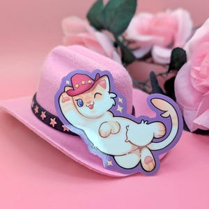 May include: A sticker of a cartoon cat wearing a pink cowboy hat, with a purple border, is placed on a pink cowboy hat. The cat is white with orange stripes and is sticking its tongue out. The hat has a black band with star details.