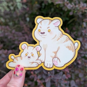 May include: A sticker featuring two cartoon bears. One bear is sitting, and the other is lying down. The bears are white with tan markings and yellow eyes. The sticker has a yellow border.
