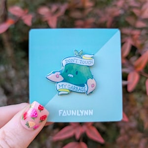 May include: A green and yellow enamel pin with a cartoon monster holding a sign that says "Don't touch my garbage". The pin is on a light blue background with the word "Faunlynn" printed below it.